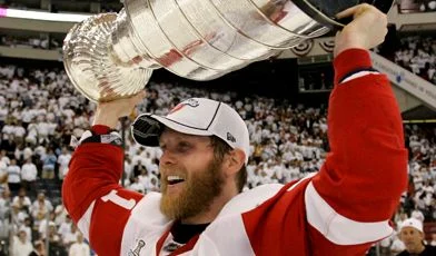 A man holding up the cup while wearing a red and white uniform.