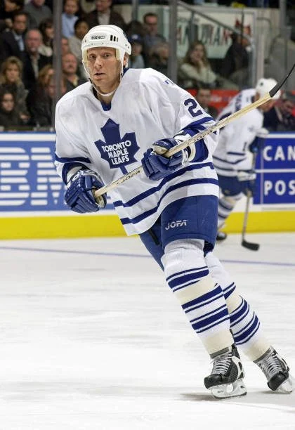 A hockey player in white and blue uniform with two sticks.